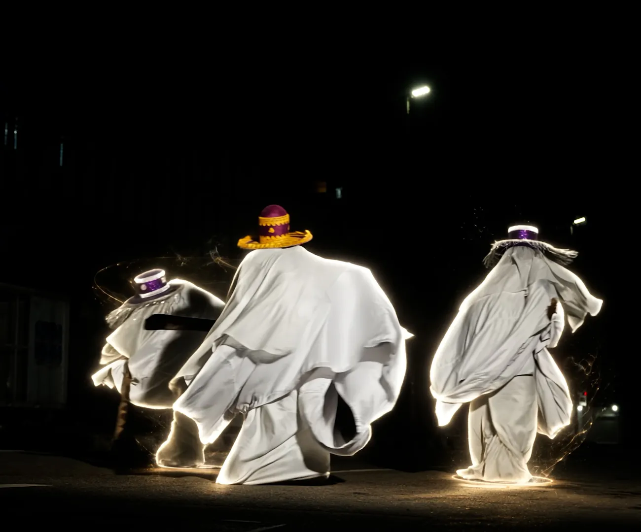 Three dancers in white robes and ceremonial hats mid-movement on a night street.